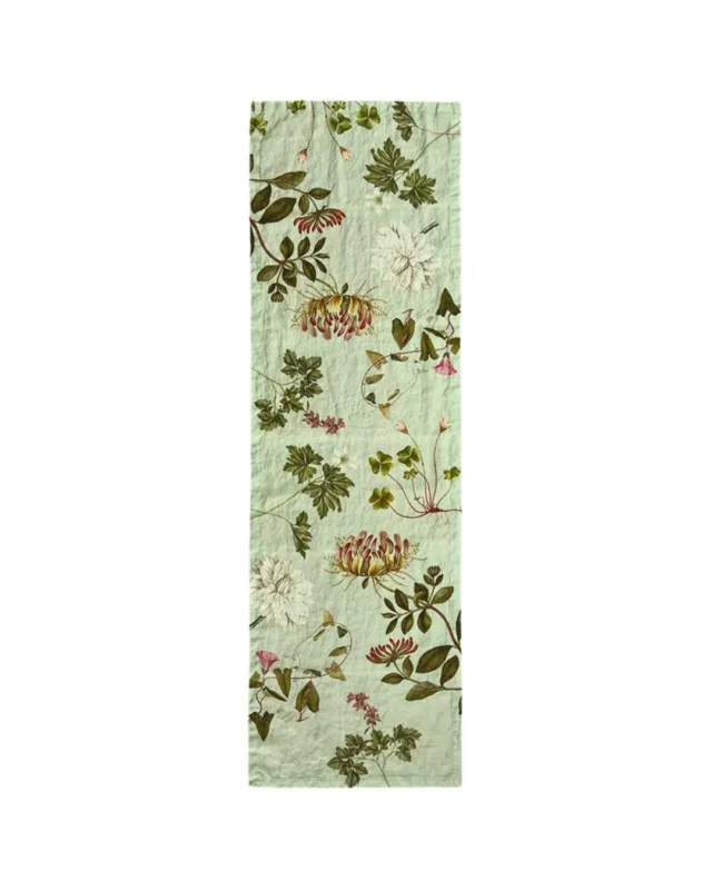 Ein schmaler Tischläufer aus Leinen mit botanischen Mustern in sanften Grüntönen, Rosa und Beige. Die natürliche, leicht strukturierte Oberfläche des Leinens unterstreicht die zarte Eleganz des "Flora Danica Atlas"-Designs von Koustrup & Co.