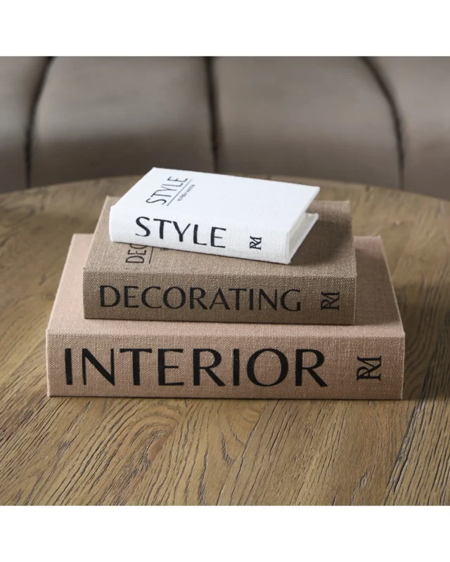 Deko Bücher "Interior Decorative Book Set" von Riviera Maison. Die 3 Bücher liegen der Größe nach geordnet aufeinander mit den Aufschriften "Interior","Decorating" und "Style" auf einem runden Holztisch im Wohnzimmer.