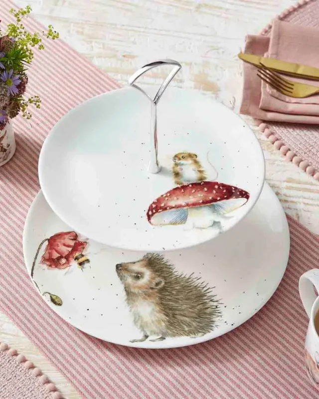 2 stöckiges Waldtier Etagere von Wrendale Designs mit Igel und Blumen Motiv auf der ersten Etage und einem Maus und Fliegenpilz Motiv auf der zweiten. Das Etagere liegt auf einem pink dekorierten/gedeckten Tisch.