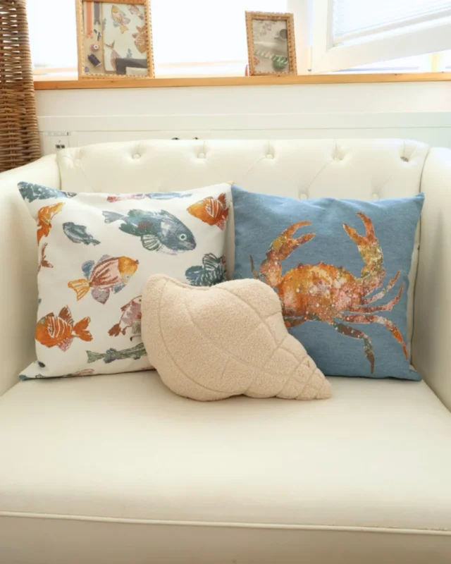 3 Maritime Kissen auf einem weißen Sofa. Zwei sind von Sander Table Home mit einem hellen Fisch Design und einem blauen Krabben Design. Das vorderste Kissen ist ein helles Muschel von Riviera Maison. Im Hintergrund sind Bilder zu sehen.