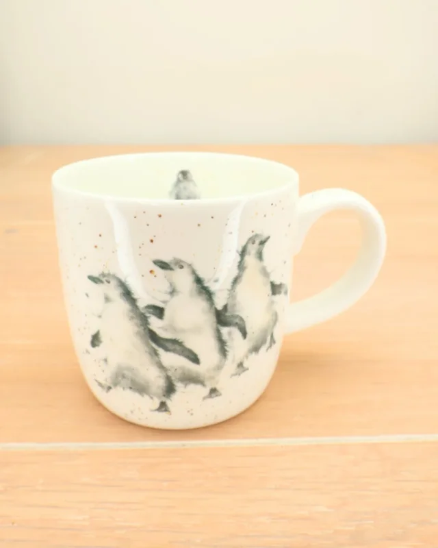 Die Pinguin Tasse "Out on the Town" von Wrendale Designs steht auf einem Holztisch. Das Motiv zeigt drei Pinguine auf hellem Grund.